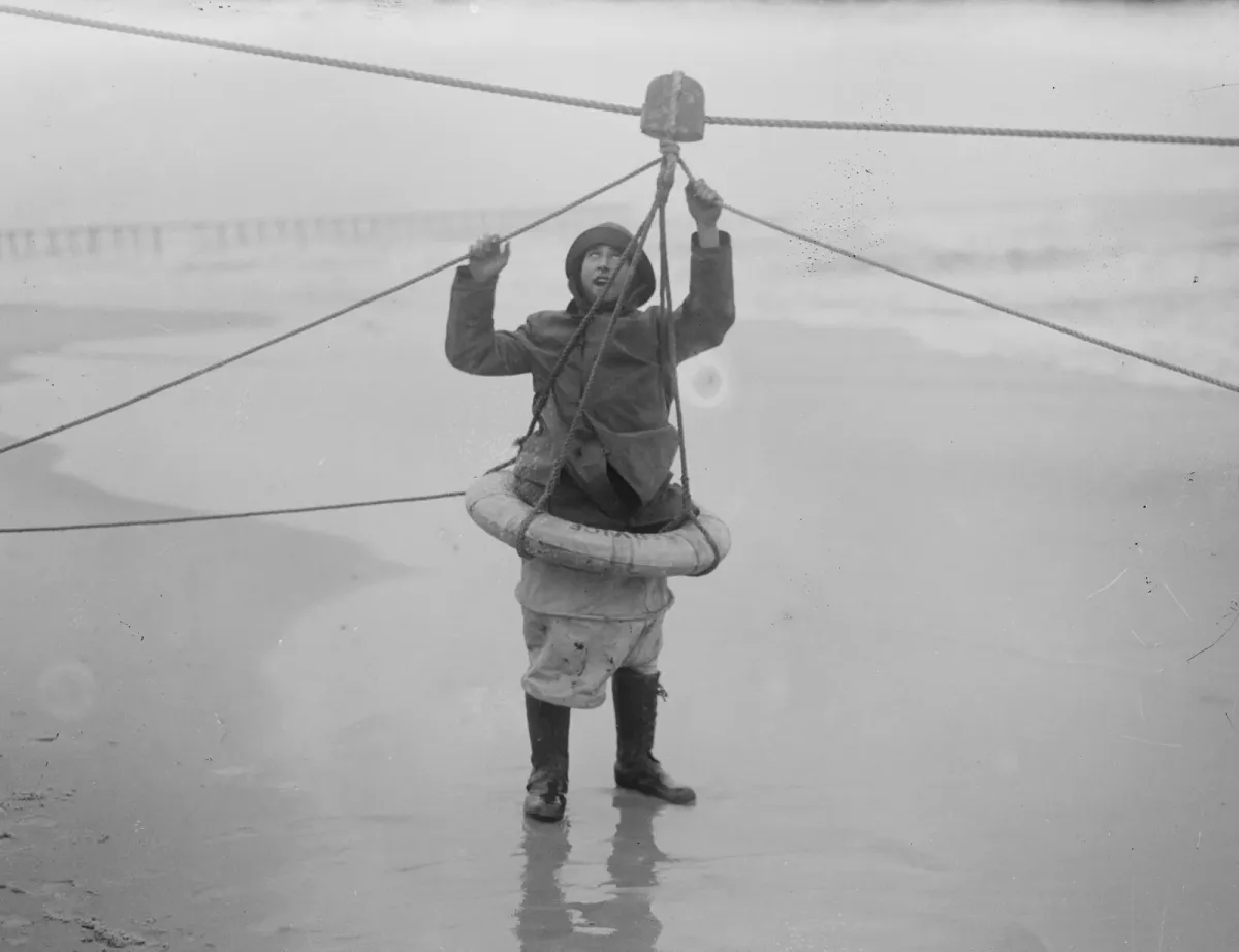 A man hanging in a breeches buoy on a rope above a beach