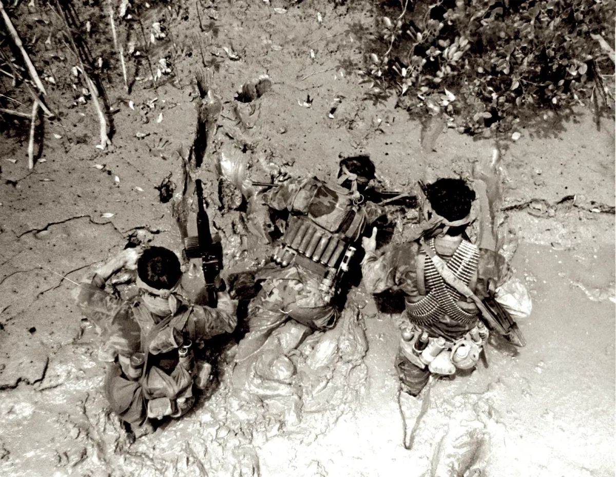 Three SEALs wading through deep mud under trees, one carrying a machine gun with belts of ammunition